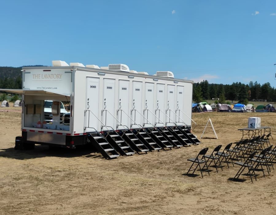 8-station shower trailer with its outside hand wash bank open on a dirt field, crew tents and pine trees behind it and folding chairs set out in front