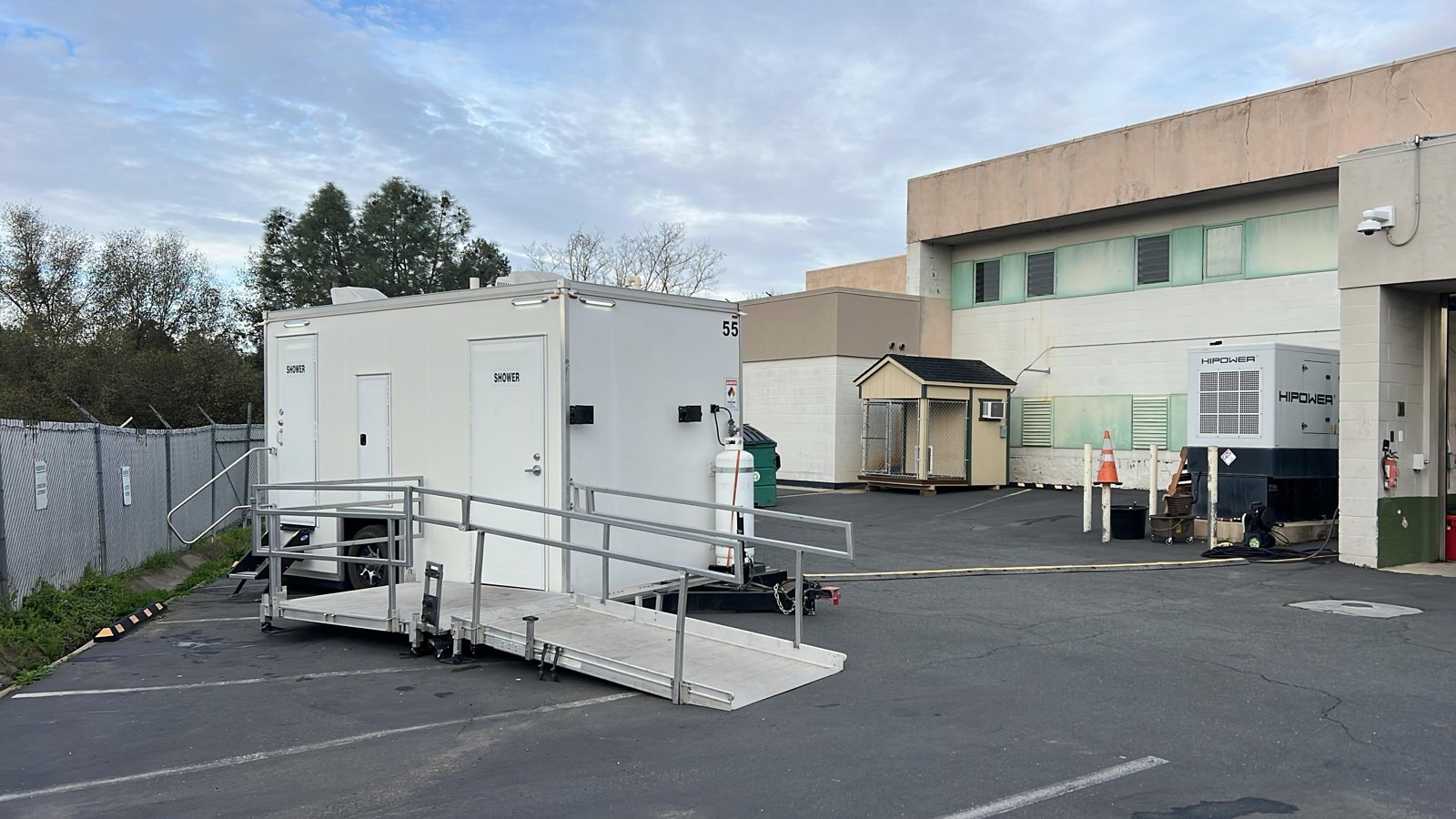 ADA plus two shower trailer with its ramp and rails deployed in the paved service yard of an industrial building next to a generator