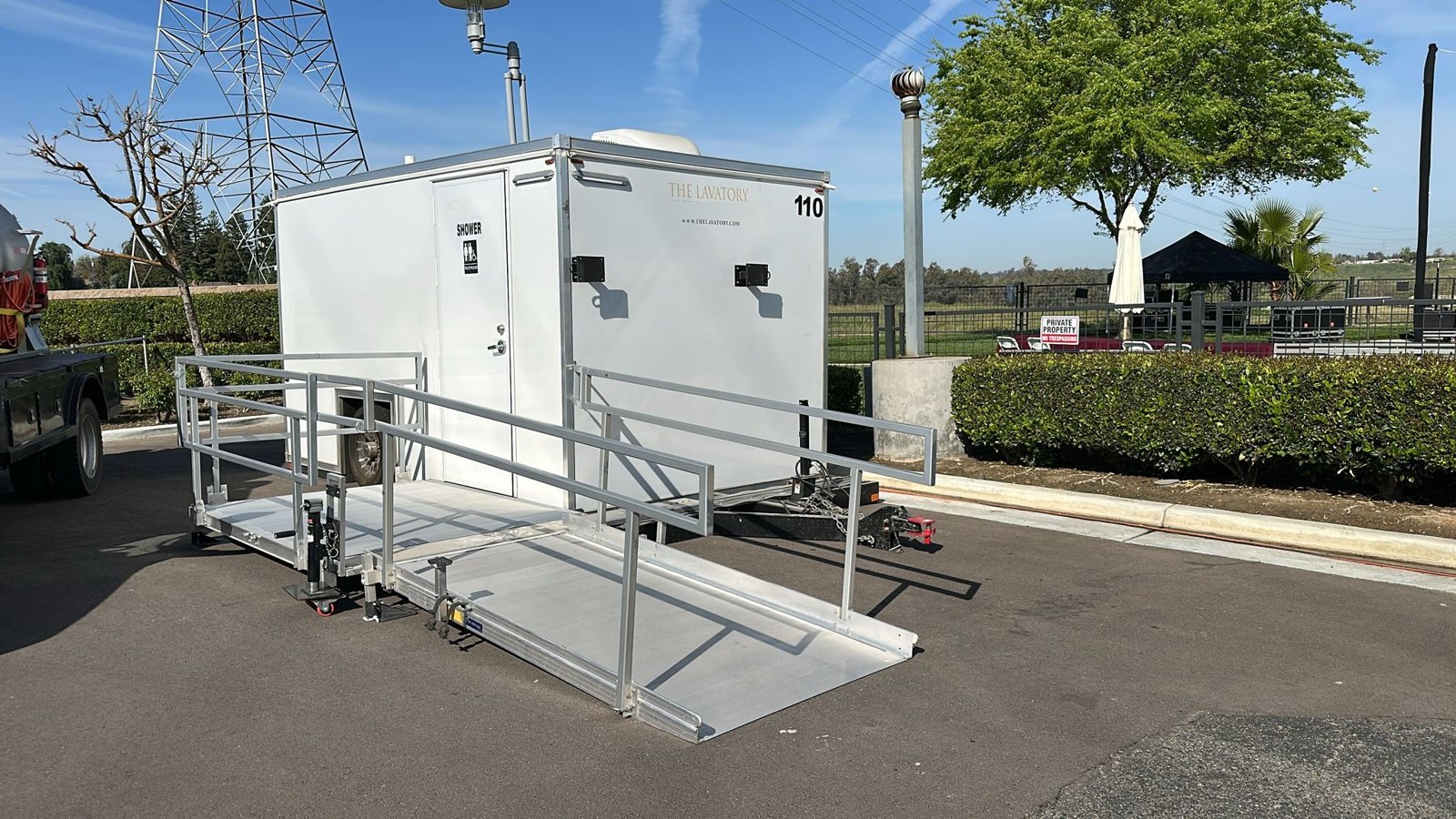 Single-station ADA shower and restroom combo trailer with a long aluminum ramp set up on a paved lot beside a hedge