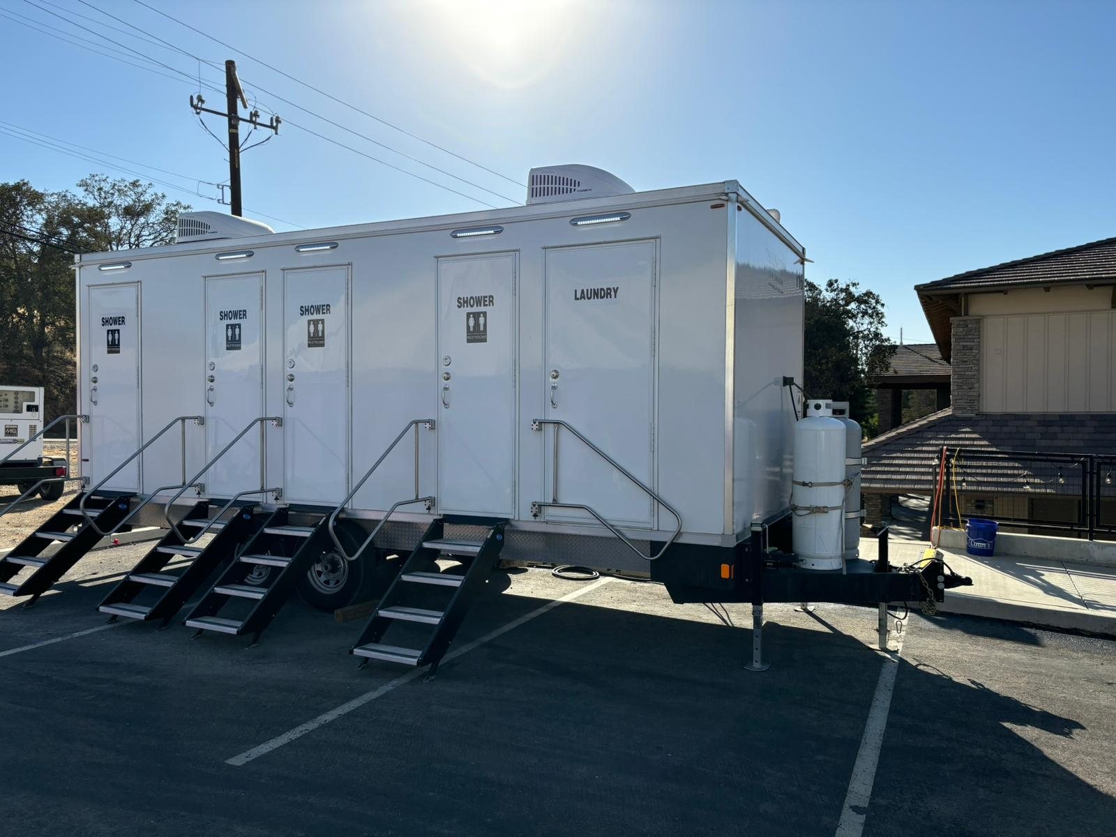 4-station shower, restroom and laundry trailer with black stair units on a paved lot below dry hills, a portable generator behind it