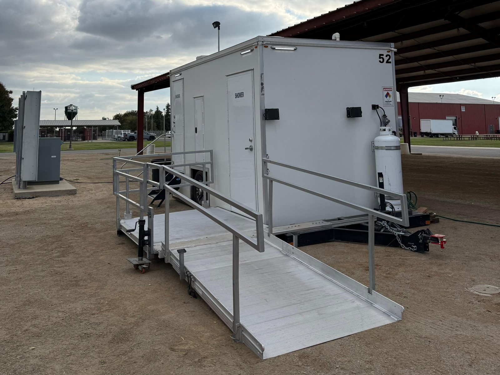 ADA plus two shower trailer with a railed aluminum ramp on a dirt lot beside a covered fairground pavilion under cloudy skies