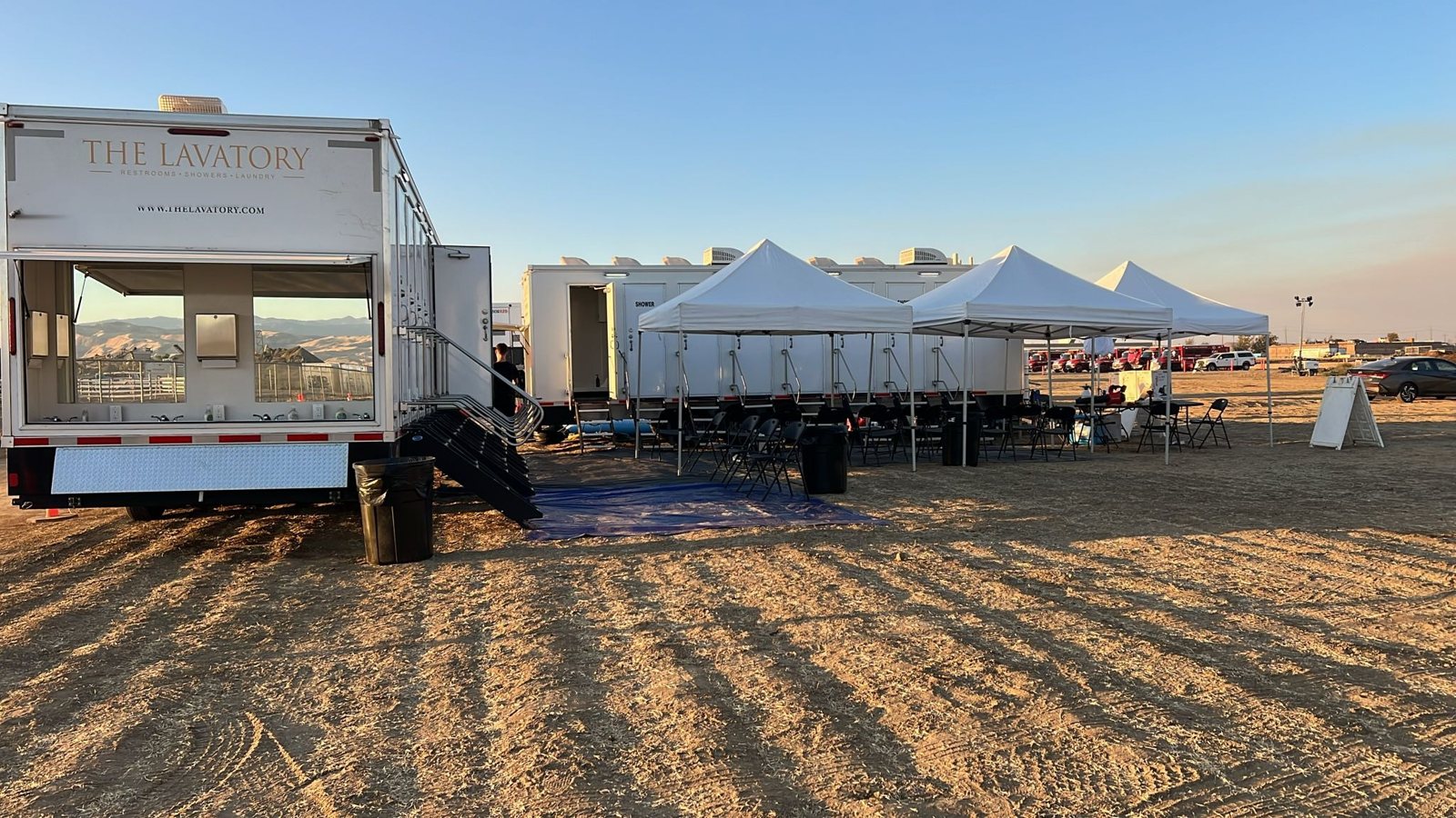 8-station shower trailer with its outside hand wash bank open beside a second shower trailer and white canopies on a dirt base camp lot, fire engines parked in the distance