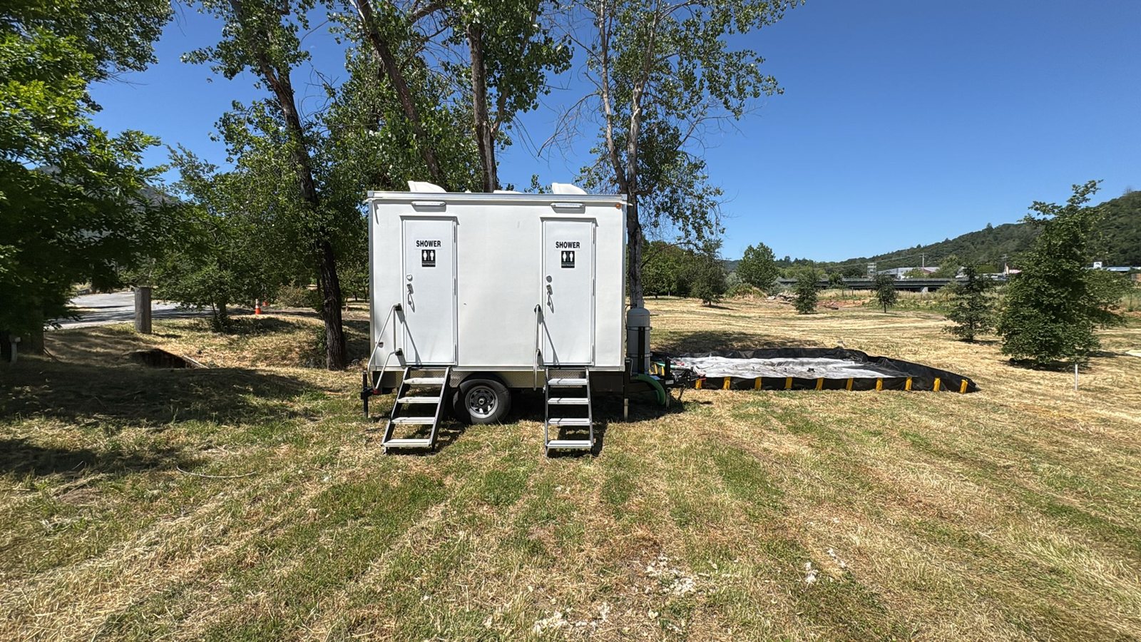 Two-door shower trailer on a mowed field under shade trees with a black spill containment berm set out beside it and green hills behind