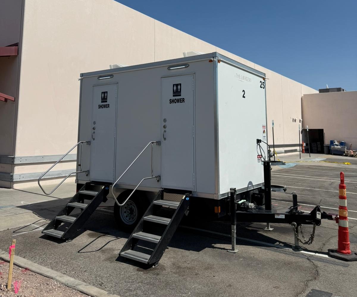 Two-room shower and restroom trailer with folding stairs parked on a paved lot beside a large commercial building, an orange cone at the tongue