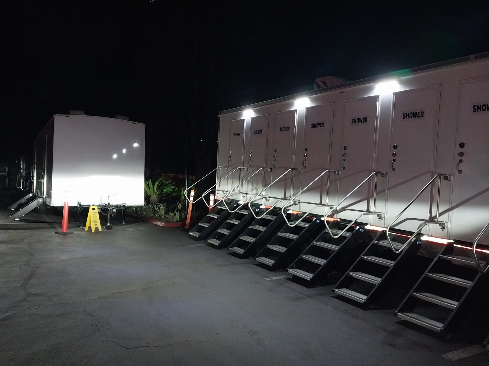 8-station shower trailer lit up at night with its door lights on, a row of stair units with rails along a paved lot and a second trailer parked behind it