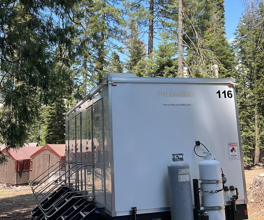 Multi-room shower trailer with stair units and propane cylinders on a dirt pad under tall pines, small red-roofed cabins behind it
