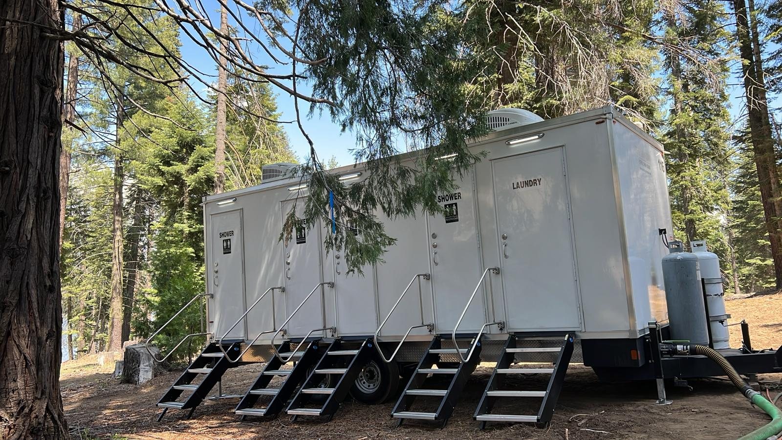 4-station shower, restroom and laundry combo trailer with five stair units set on a pine-needle clearing in a conifer forest, a sewer hose run from the tongue
