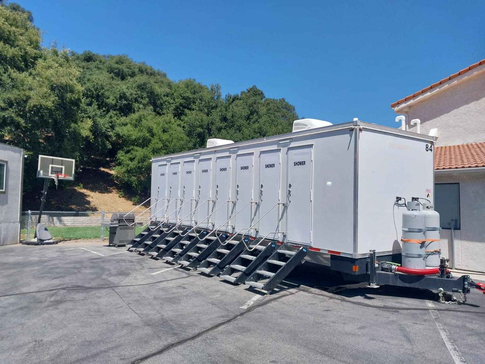 8-station private shower trailer with eight doors and eight stair units parked on a paved lot below an oak-covered hillside, next to a stucco building with a red tile roof