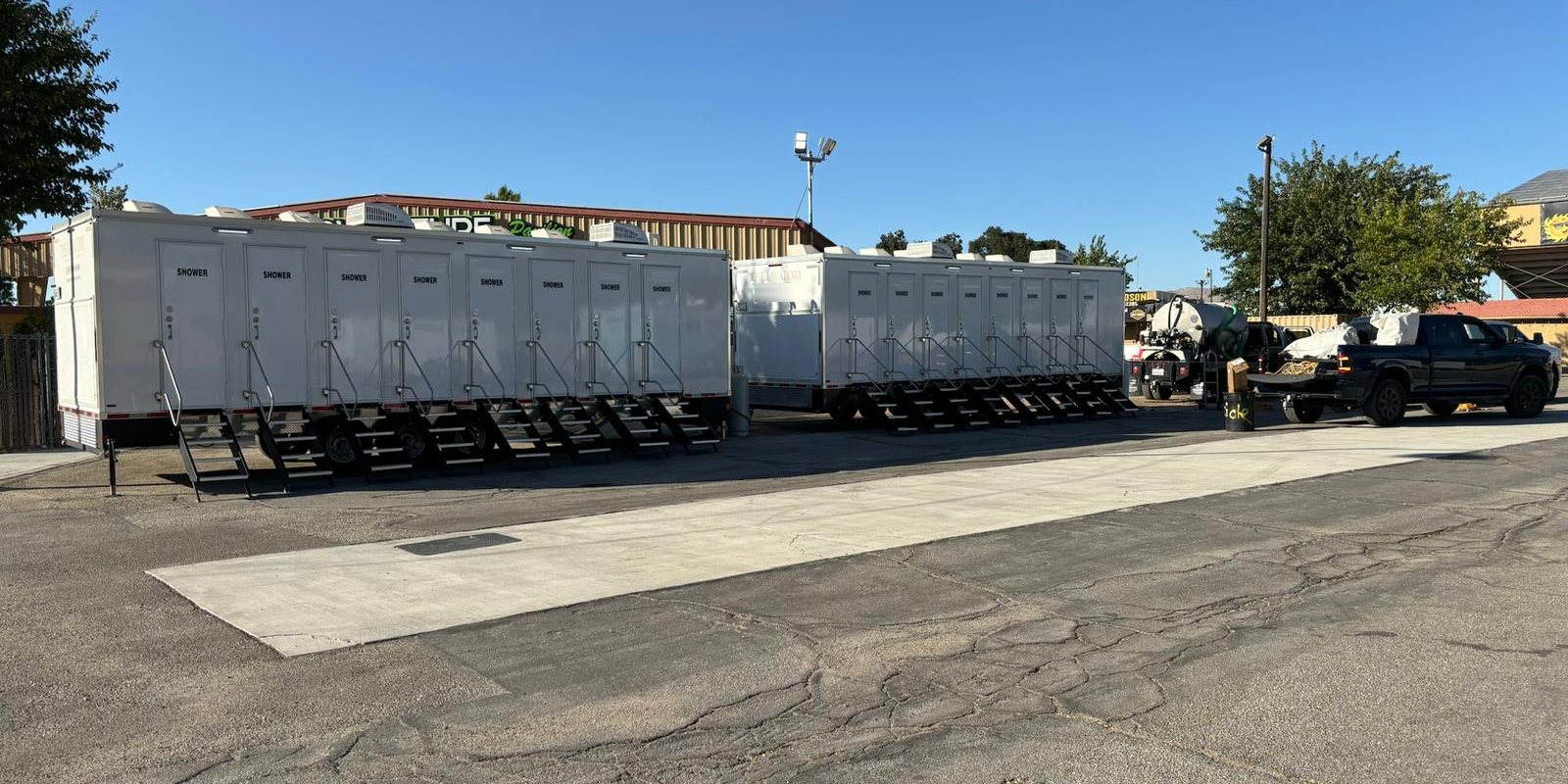 Two 8-station shower trailers parked end to end on a paved event lot with their stair units set, a service truck and a pickup parked beside them