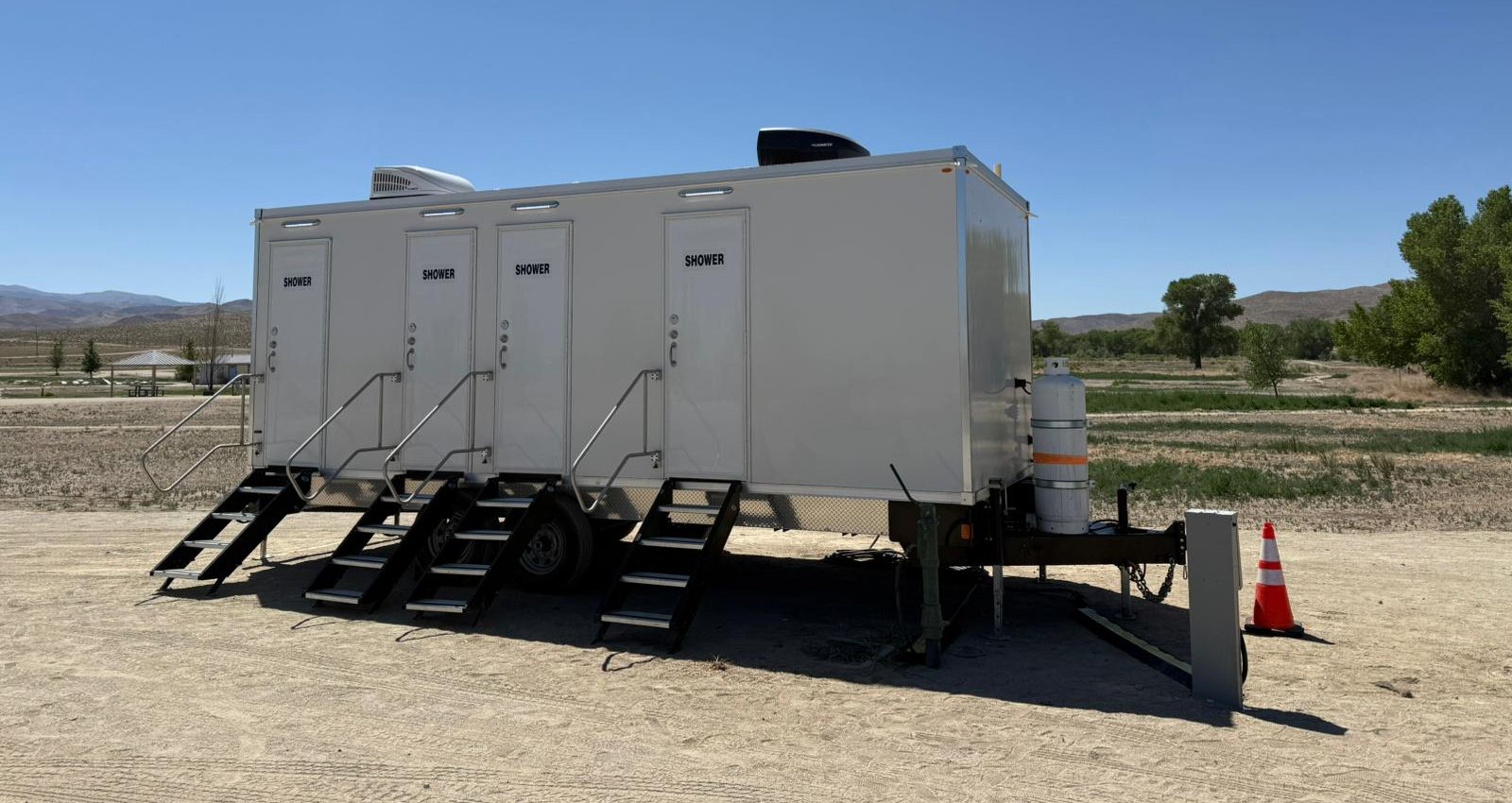 Four-door shower trailer with its stair units set on a sandy gravel yard, a power pedestal and a traffic cone at the tongue, brown hills and cottonwood trees behind it