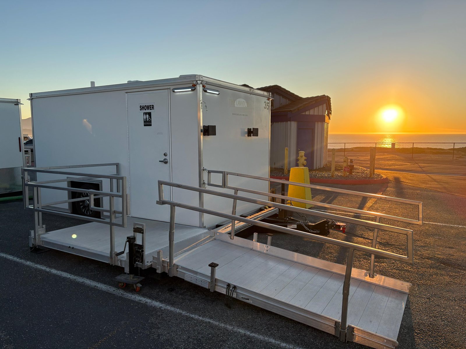 ADA plus two shower trailer with a railed aluminum ramp in a paved coastal parking lot at sunset, the ocean behind a fence
