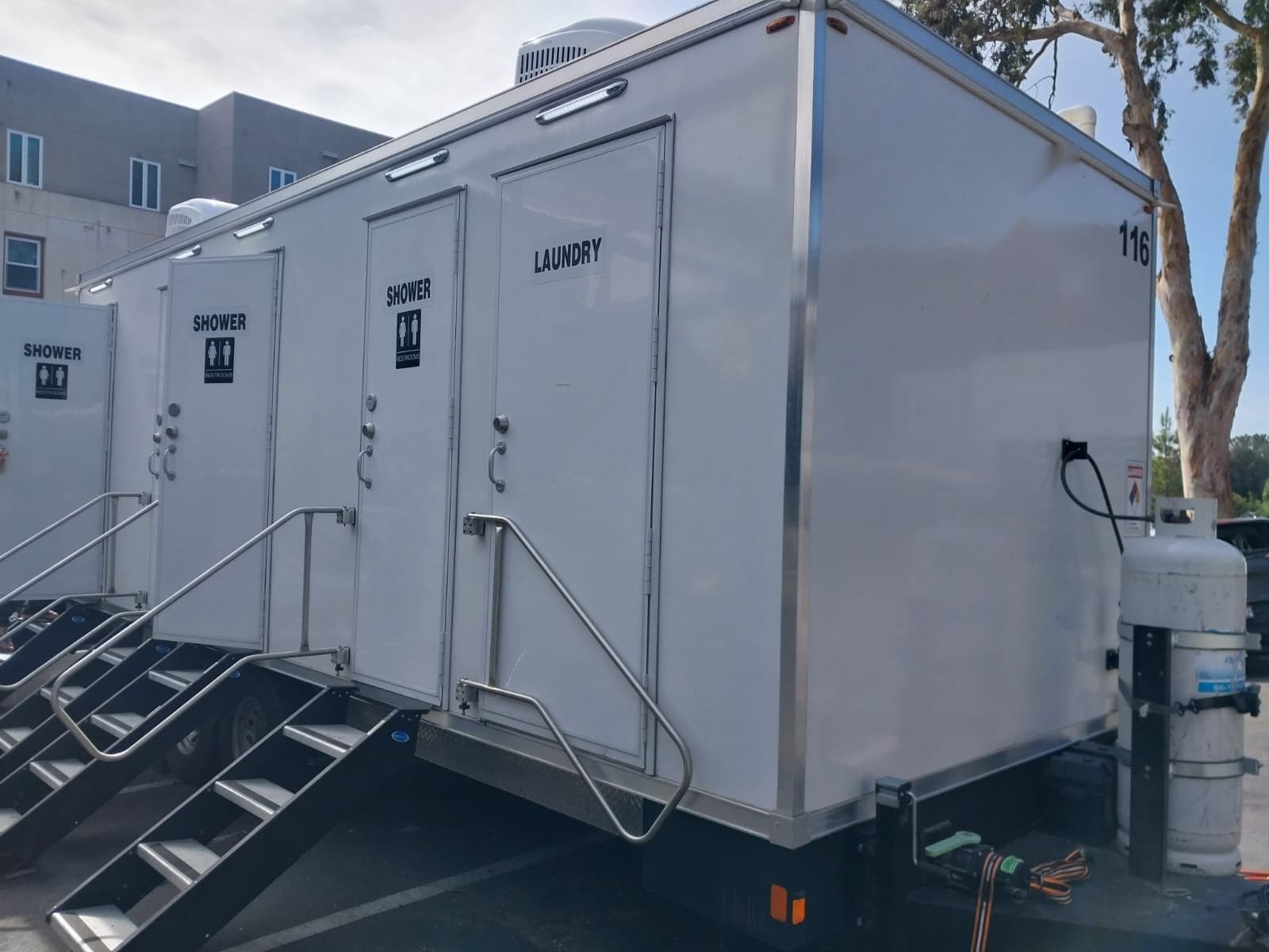 4-station shower, restroom and laundry trailer with black stair units in a paved lot beside a multi-story building
