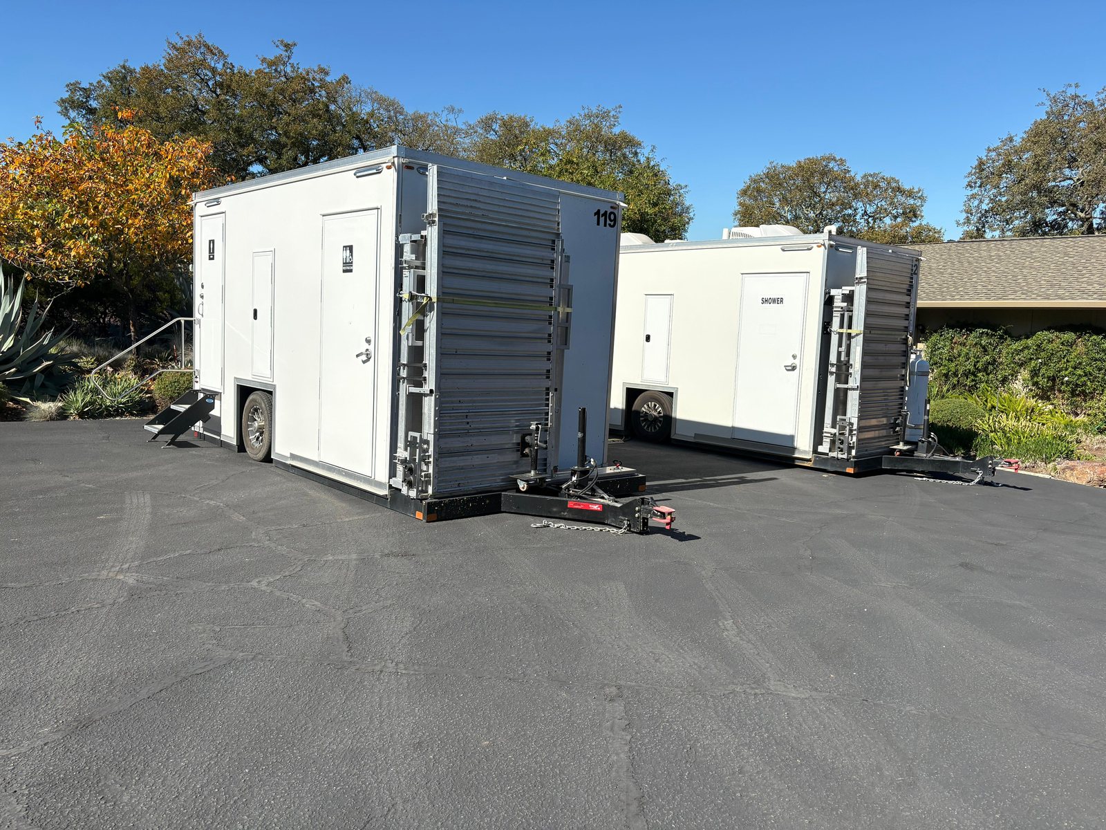 Accessible restroom trailer and a shower trailer with folded ramps parked on a paved lot beside trees and shrubs
