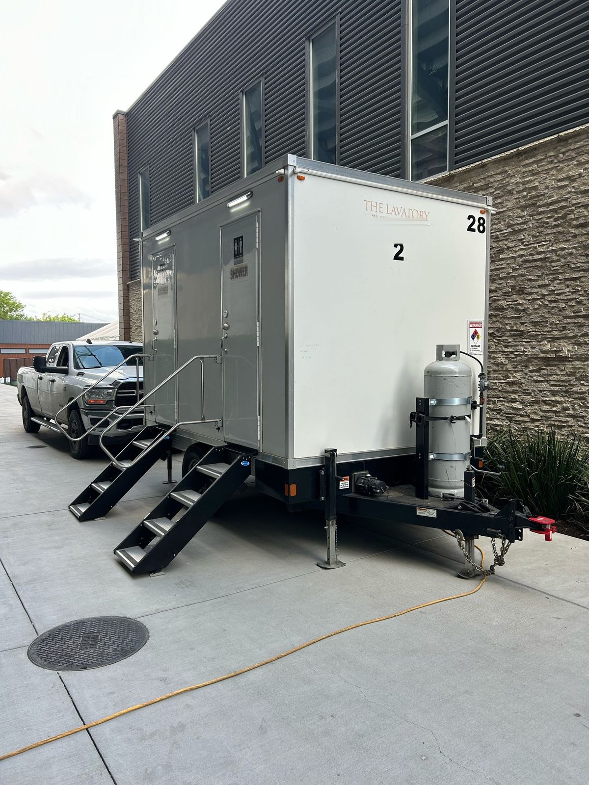 2-station shower and restroom trailer on a concrete apron beside a modern building, a pickup truck parked behind it