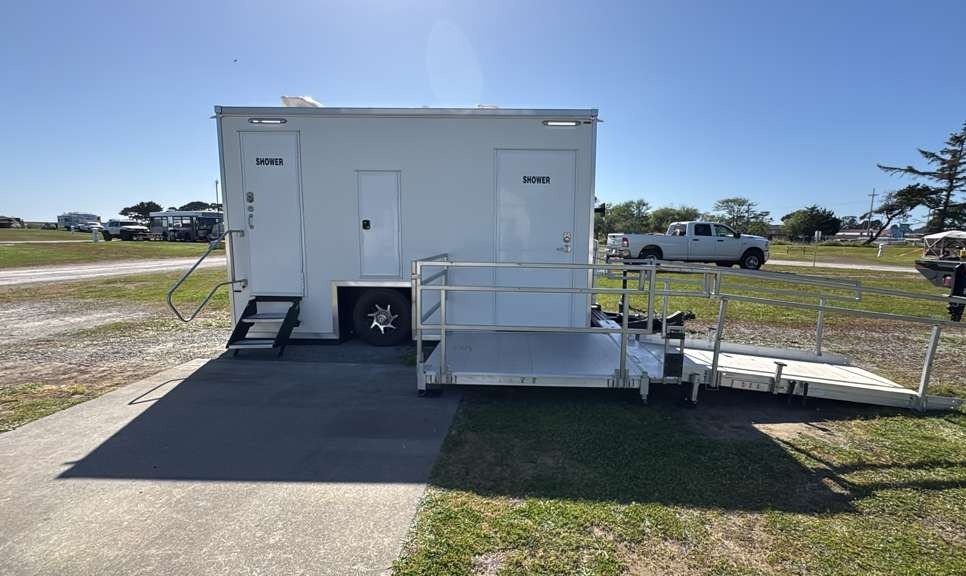 ADA + 2 shower trailer with a railed aluminum ramp and side stairs on a paved pad at the edge of a grassy coastal field