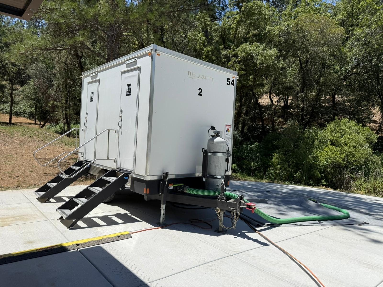 2-station shower and restroom combo trailer hooked to water and sewer hoses on a new concrete pad below an oak and pine slope