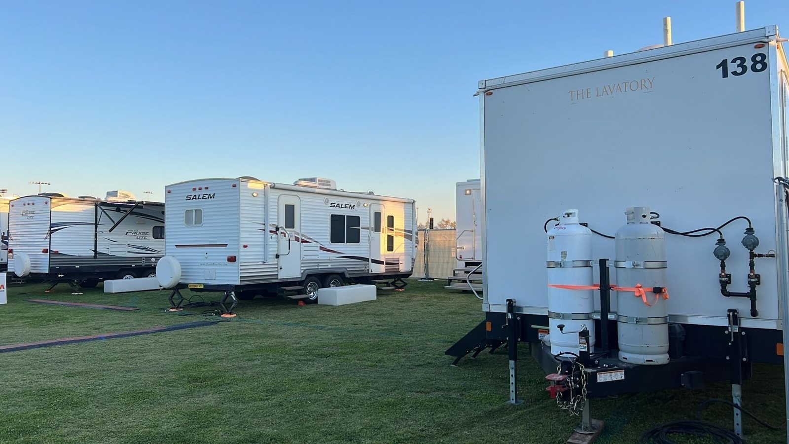 Rear of an 8-station shower trailer with propane tanks beside a row of travel trailers on a grass base camp at sunrise