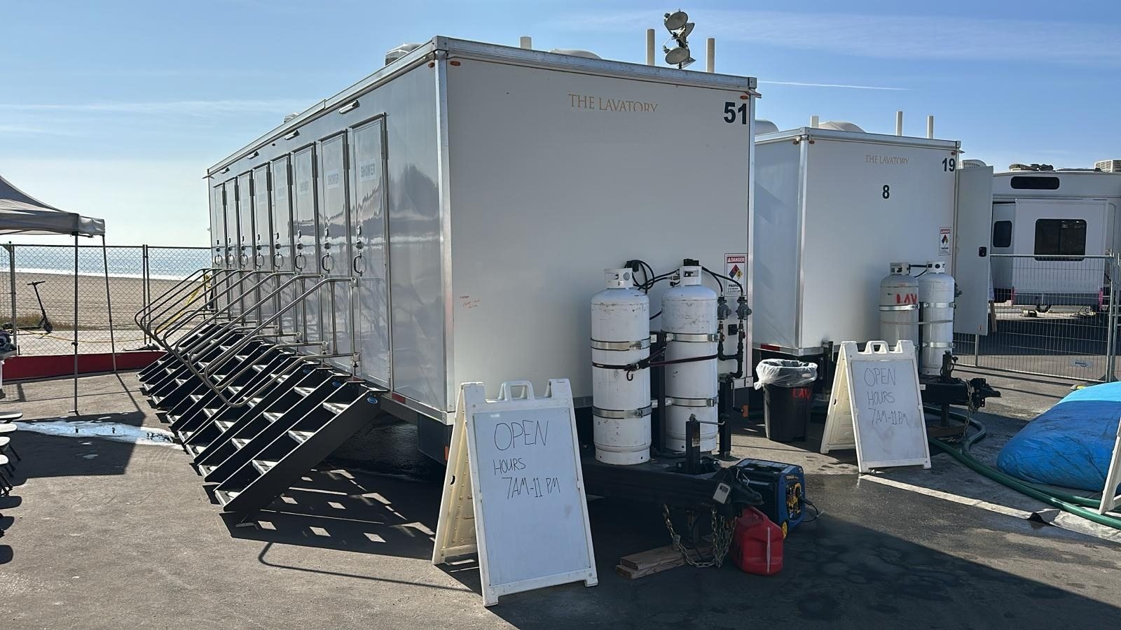 Two 8-station private shower trailers with stair units on a paved base camp lot by a beach, open-hours signs and propane tanks beside them