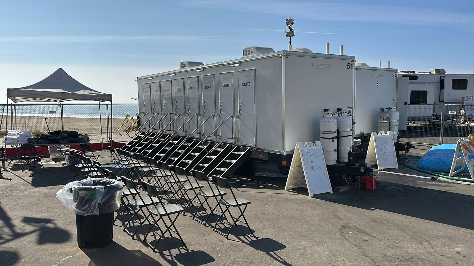 8-station private shower trailer with eight doors and eight stair units on a paved lot beside the beach, with rows of folding chairs, a pop-up canopy and two open-hours signs in front