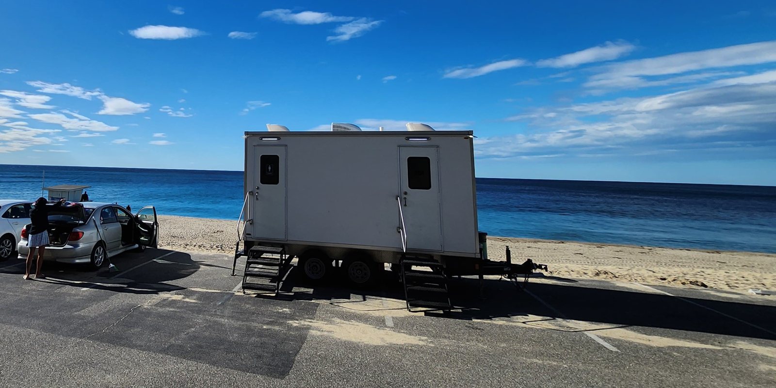 2-station shower and restroom combo trailer with two stair units on a beach parking lot facing the ocean, a beachgoer loading a car nearby