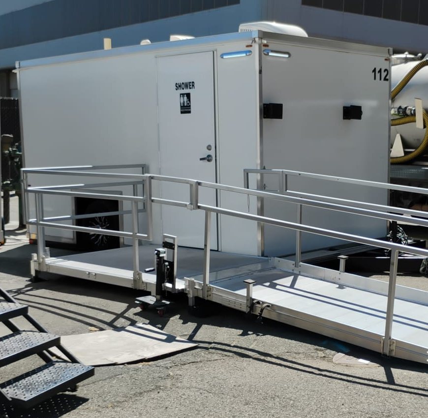 Single-station ADA shower and restroom trailer with a long ramp and handrails on a paved industrial yard, a service truck tank at its side