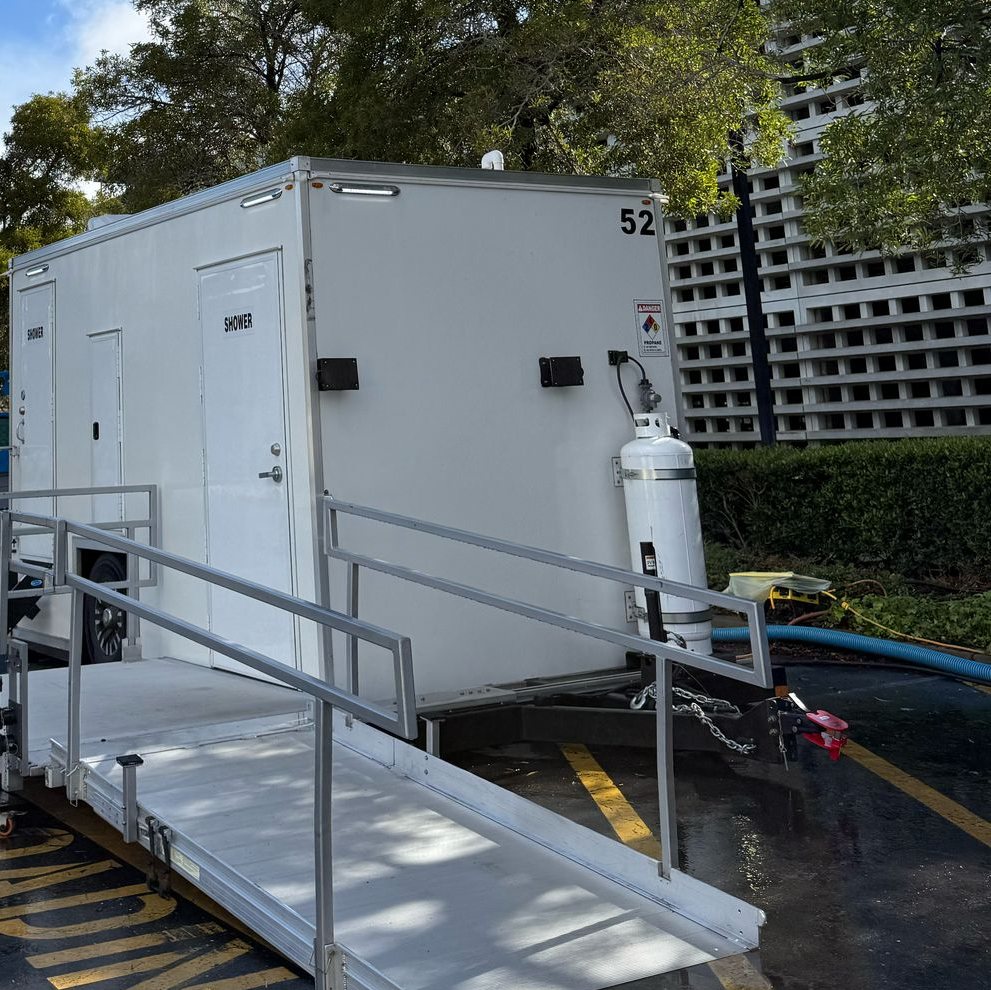 ADA plus 2 shower trailer with its aluminum ramp and handrails set in a wet paved loading zone, trees and a concrete building behind it