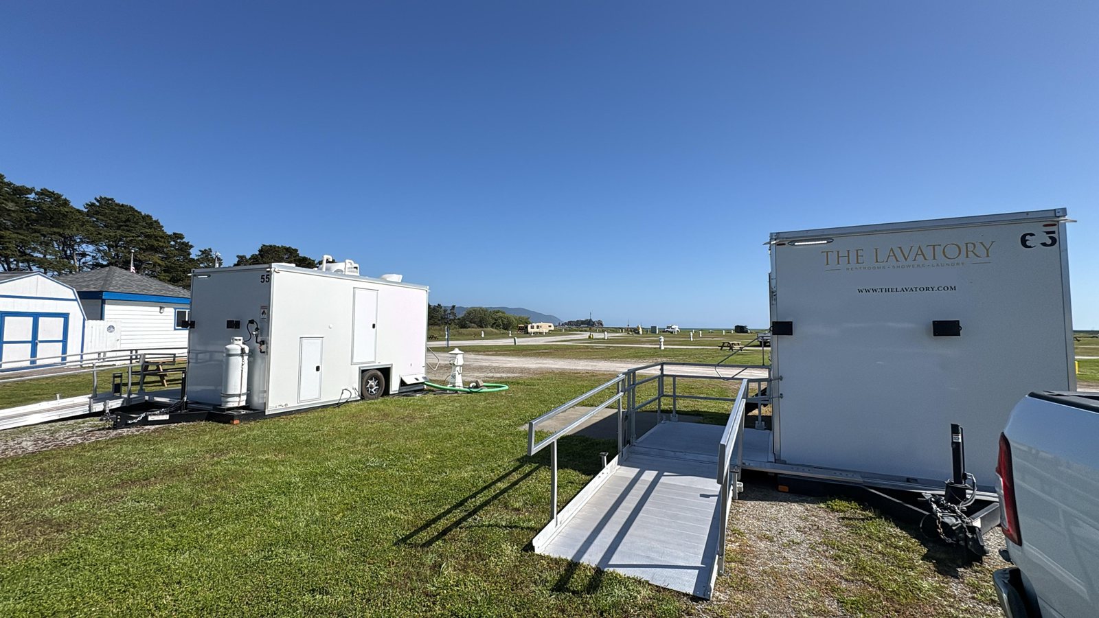 ADA shower trailer with an aluminum ramp set on a grass event field, a second trailer parked beside a small building