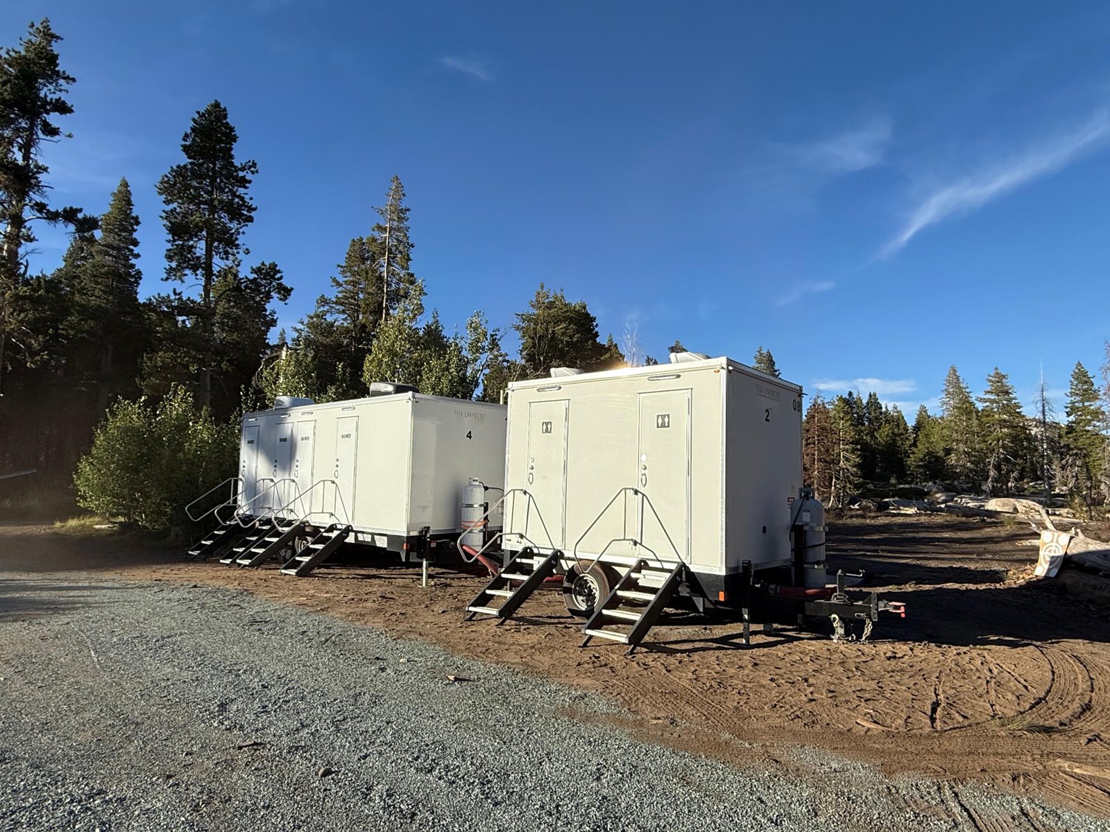 4-station shower trailer and a 2-station trailer parked side by side on a dirt pad beside a gravel road, with pine trees behind them