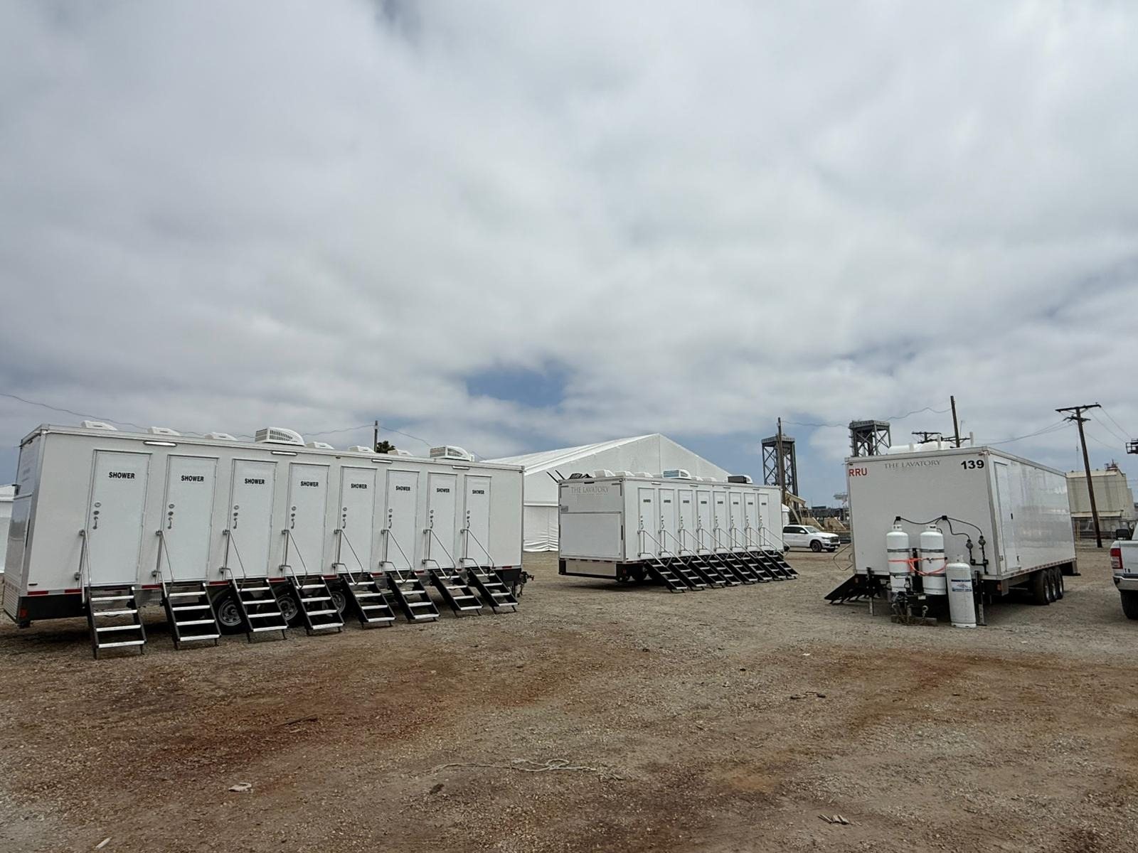 Two 8-station shower trailers and a support trailer set up on a gravel industrial yard next to a large white tent and steel towers