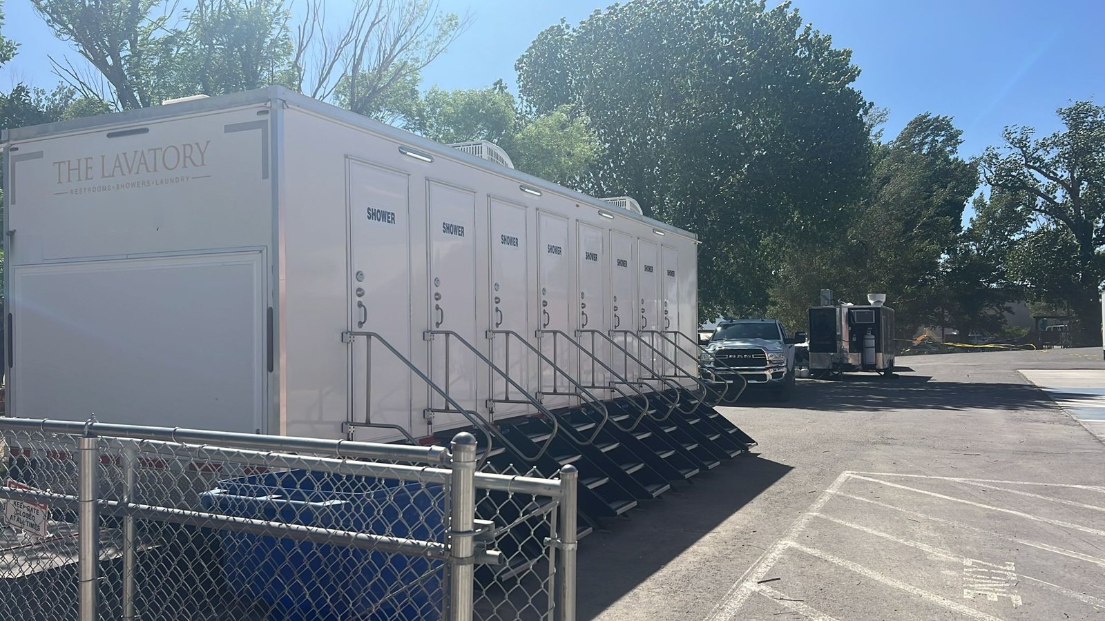8-station private shower trailer with a stair at each of its eight SHOWER doors, parked on a paved lot under big shade trees beside a chain link fence