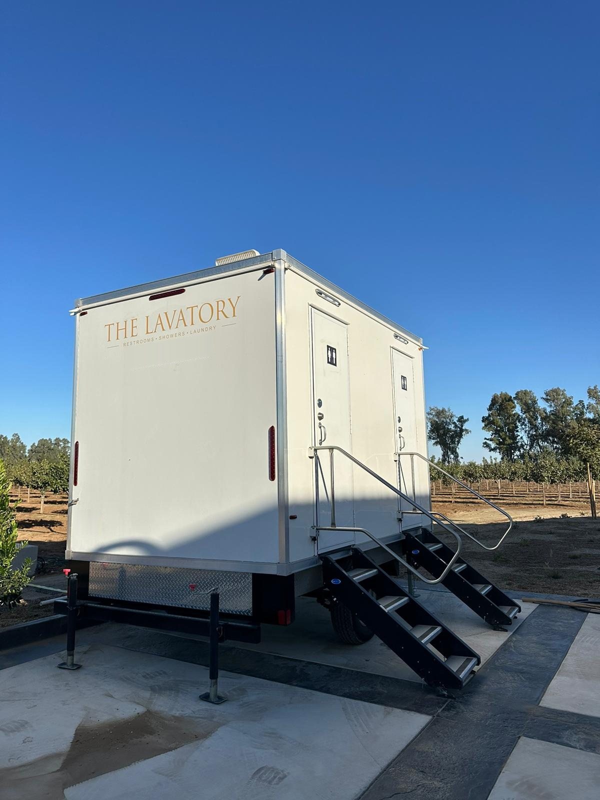 2-station shower and restroom combo trailer with two stairs, parked on a concrete pad at the edge of a young orchard