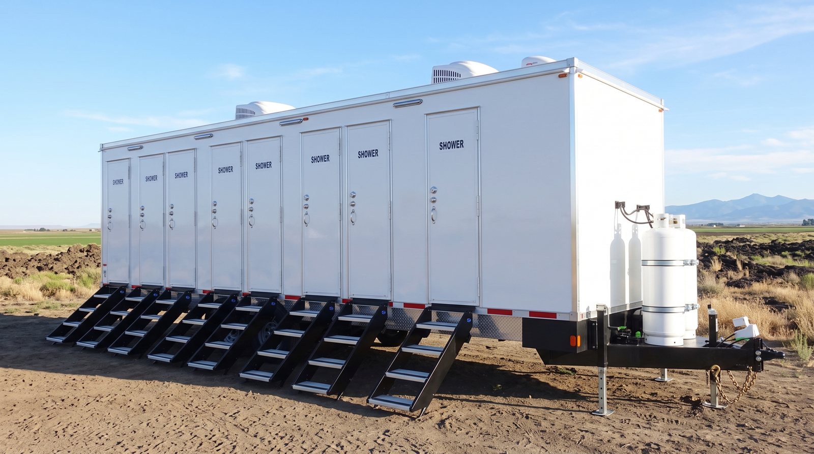 Private 8-station shower trailer on a dirt pad at the edge of farm fields with mountains on the horizon, the kind of site found around Idaho Falls, ID