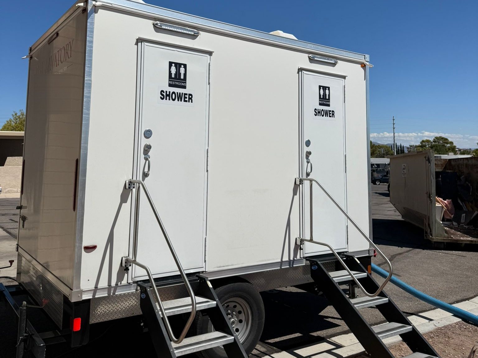 2-station shower and restroom combo with two steel stairs, set on a paved lot next to a roll-off bin under a clear blue sky