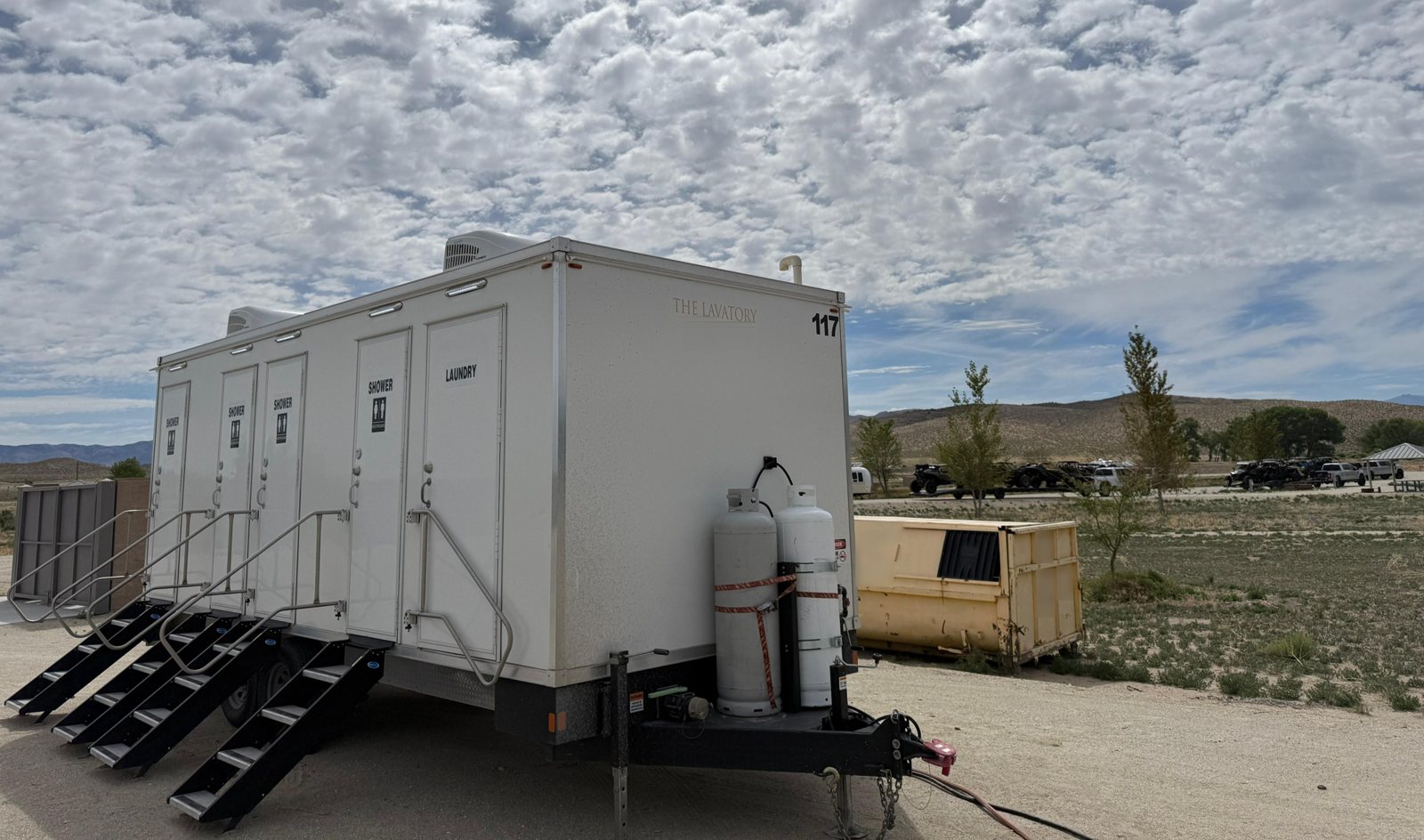 4-station shower, restroom and laundry combo trailer with four stairs and two propane tanks, parked on a dirt pad in open high desert with sagebrush hills under a cloudy sky