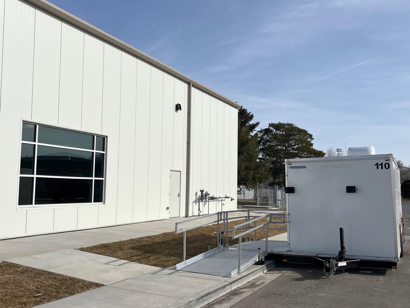 Single-station ADA shower and restroom combo with an aluminum ramp and rails, parked beside a new white metal industrial building