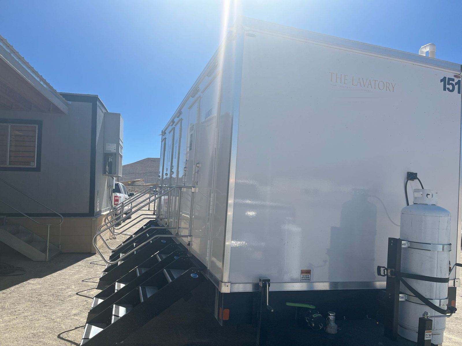 Shower, restroom and laundry combo trailer with railed stairs on a dirt jobsite yard next to a modular site office, a pickup and a hillside behind