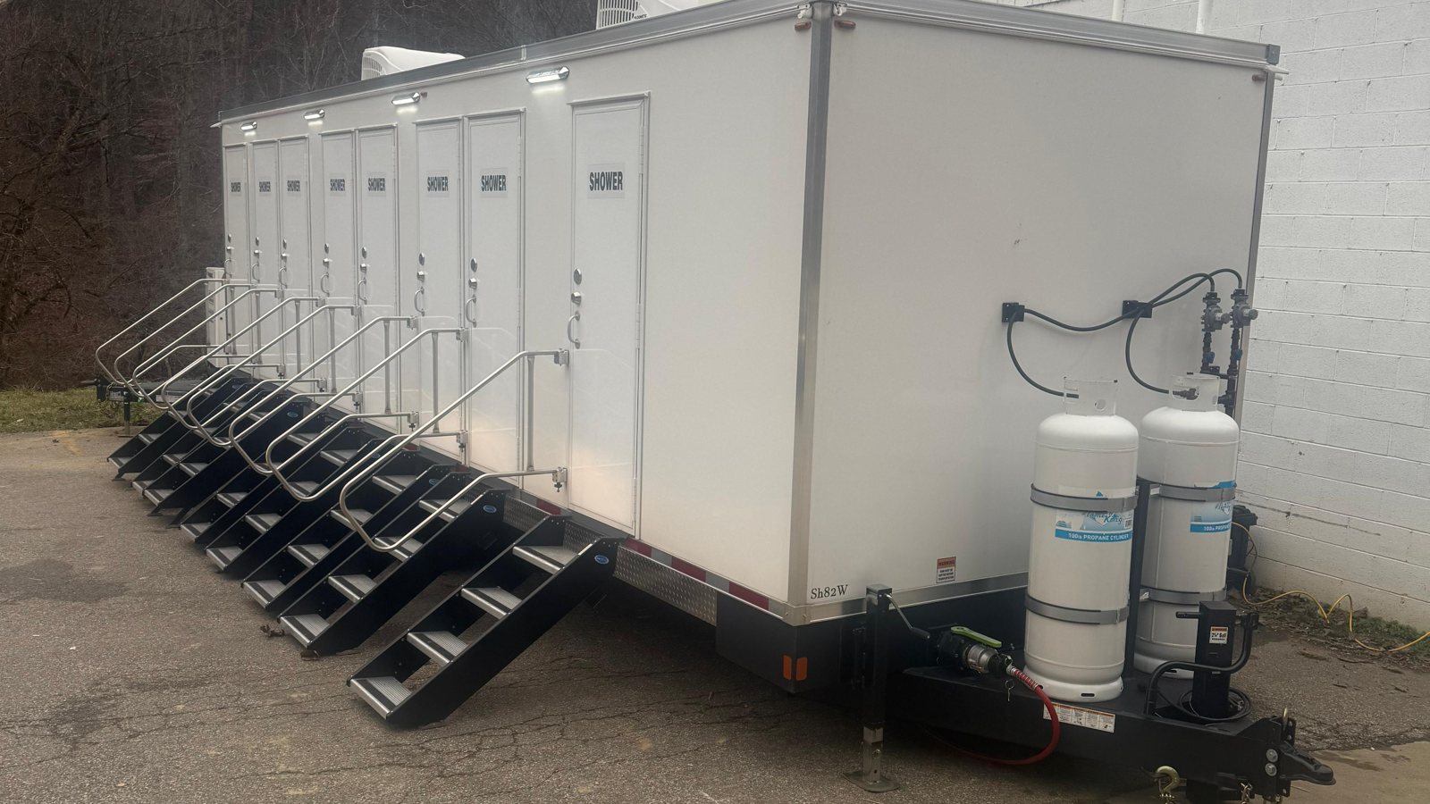 8-station shower trailer with eight doors and eight railed stair units parked beside a white block building, two propane tanks on the tongue