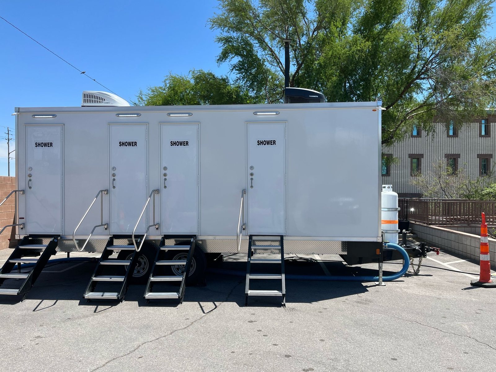 Private shower trailer with four marked shower doors and folding stair units, parked level on a paved lot under shade trees
