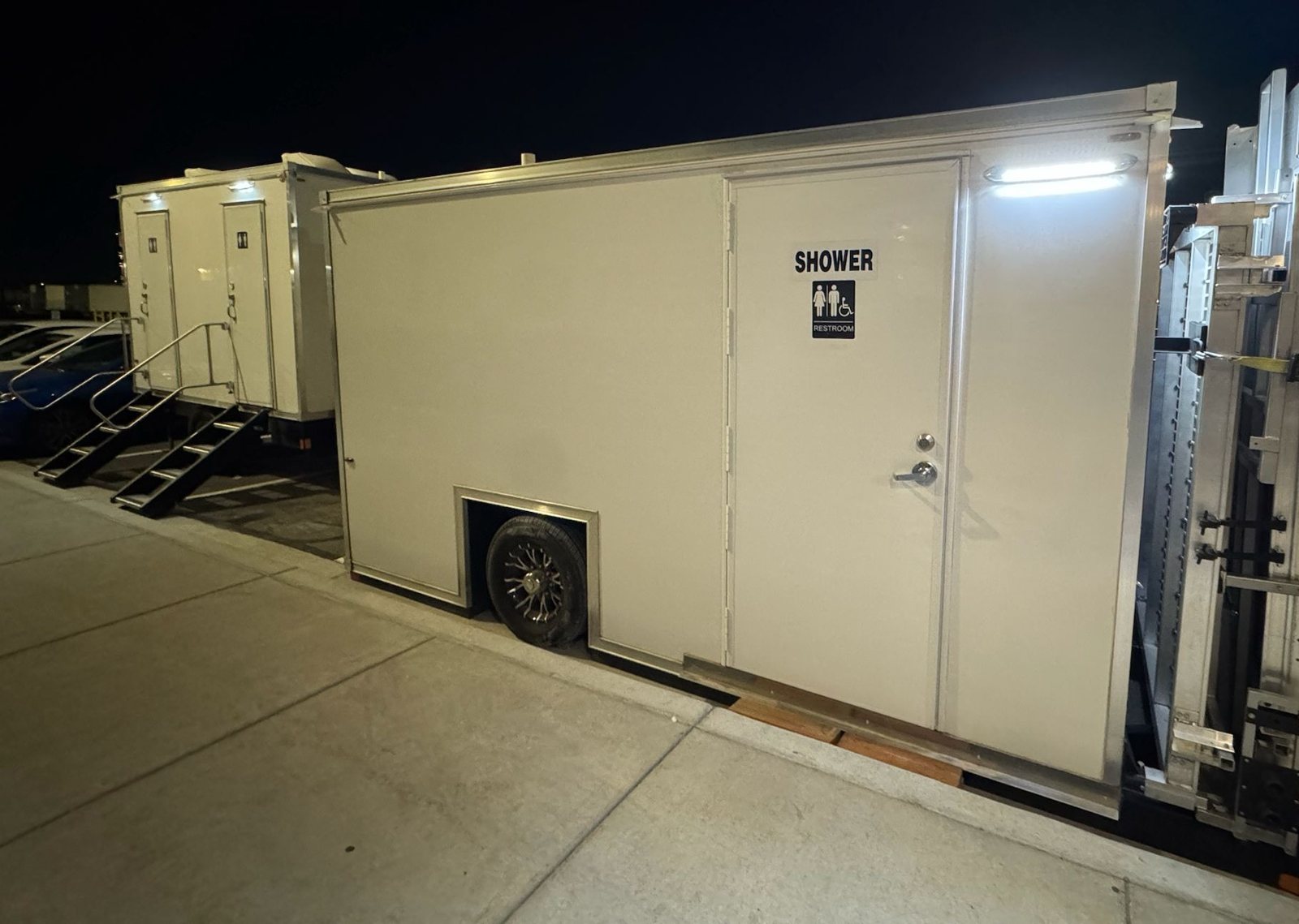 Single-station ADA shower and restroom combo lit up at night beside a curb, its folded ramp on the end and a restroom trailer behind