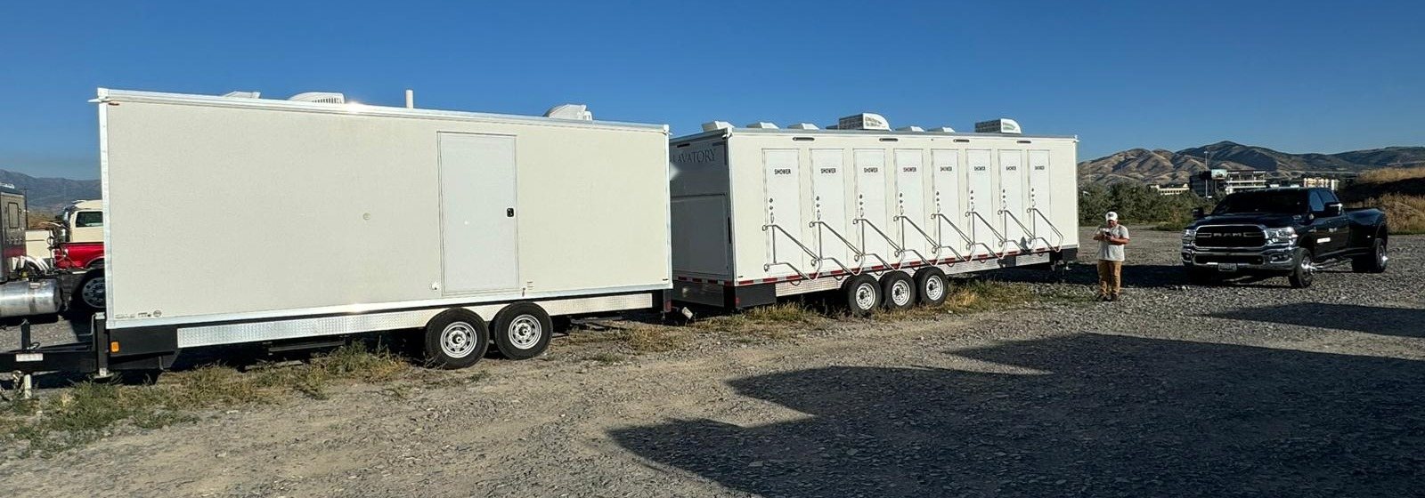 8-station shower trailer parked on a gravel yard beside a large white support trailer, a crew member and a pickup truck nearby, foothills behind