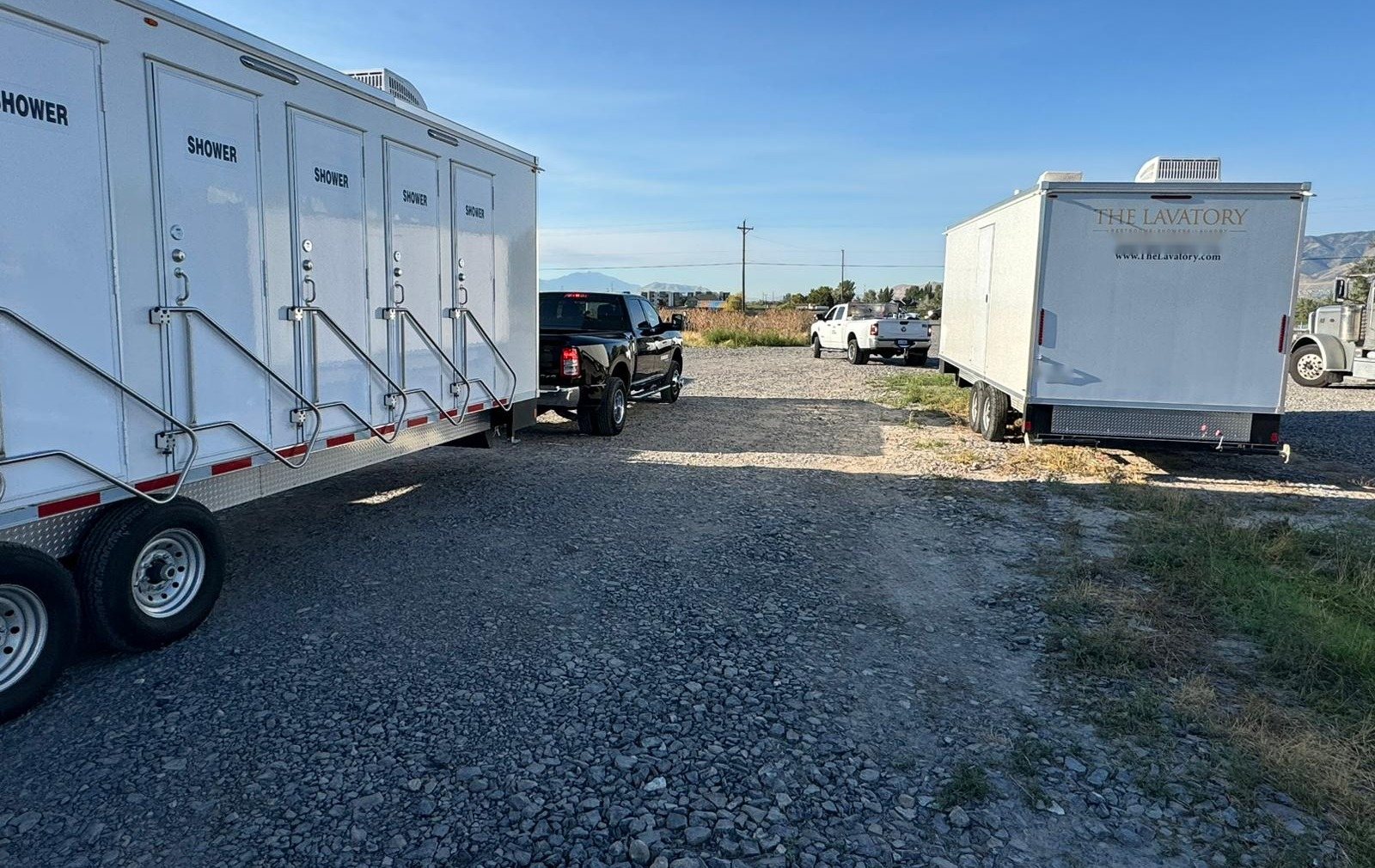 Private shower trailer doors with handrails on a gravel staging yard, a support trailer and work trucks parked across the lot
