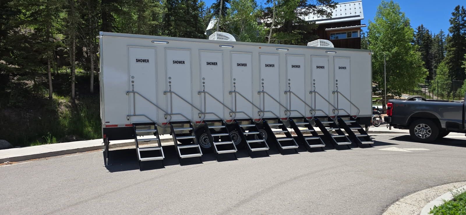 8-station private shower trailer with eight doors and eight stair units set level on a paved lot, pine trees and a lodge behind it