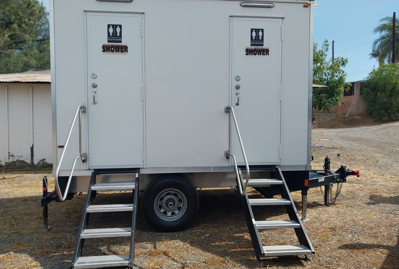 2-station shower and restroom combo trailer with a stair at each door, parked on a dry dirt yard beside a white shed and palm trees