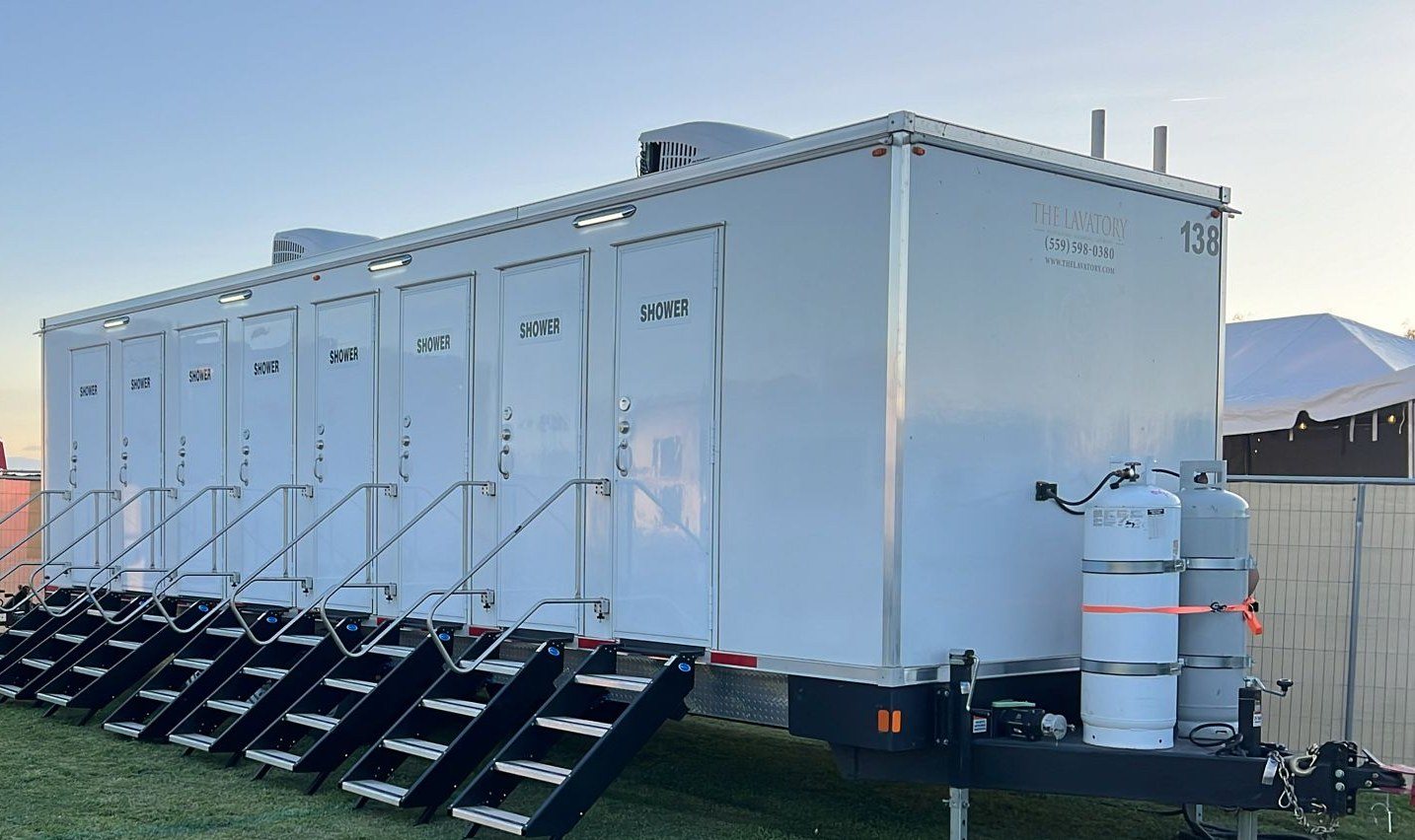 8-station private shower trailer with a stair at each of its eight doors, set up on a grass field at dusk beside a white event tent