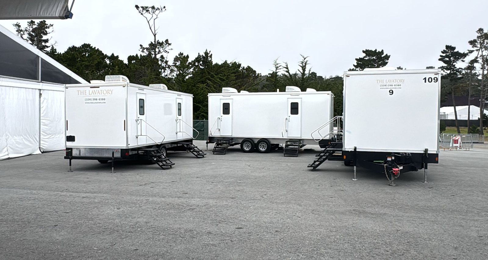 Three Lavatory trailers with their stairs set, parked on a paved lot next to large white tents and a row of trees