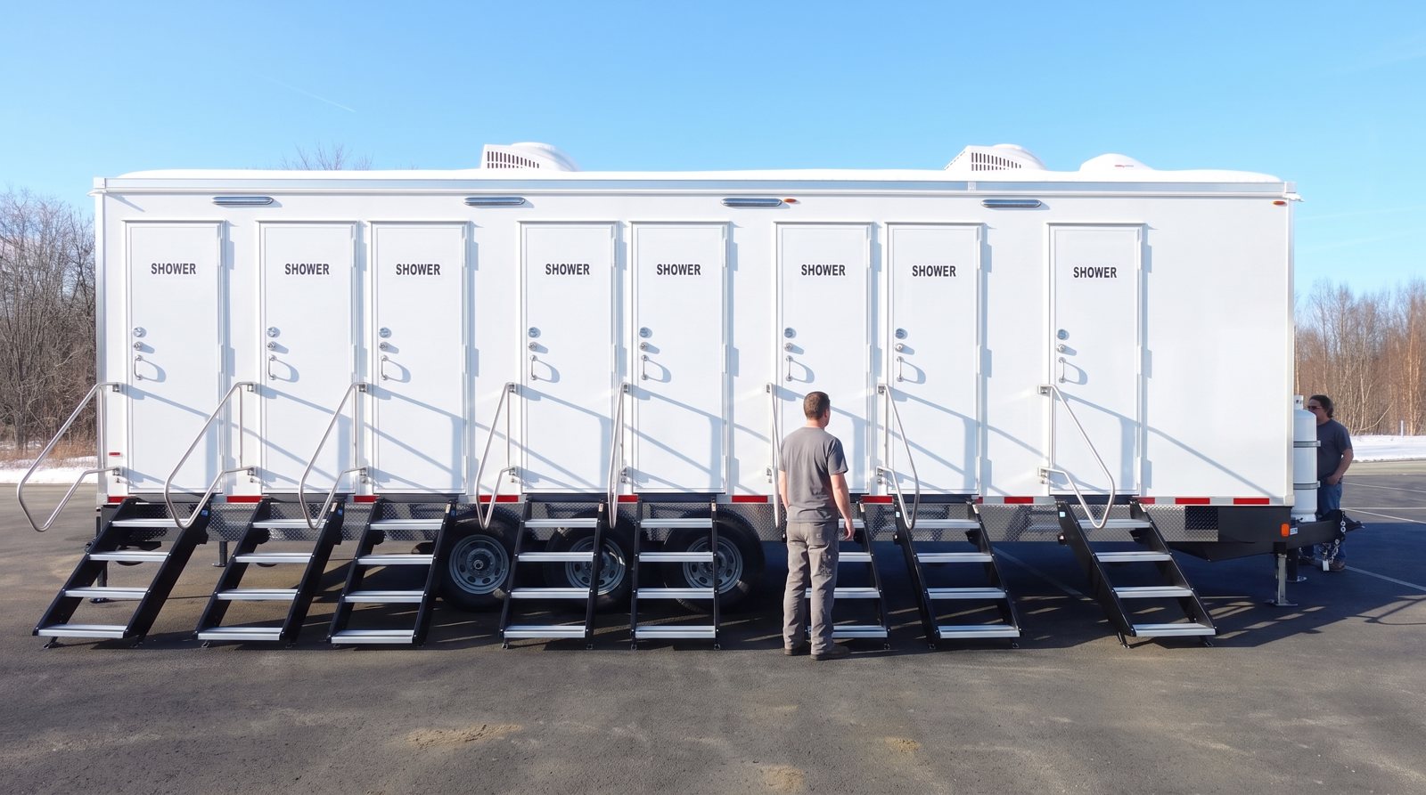 8-station shower trailer on a paved lot with a stair at each of its eight doors and two crew members beside it