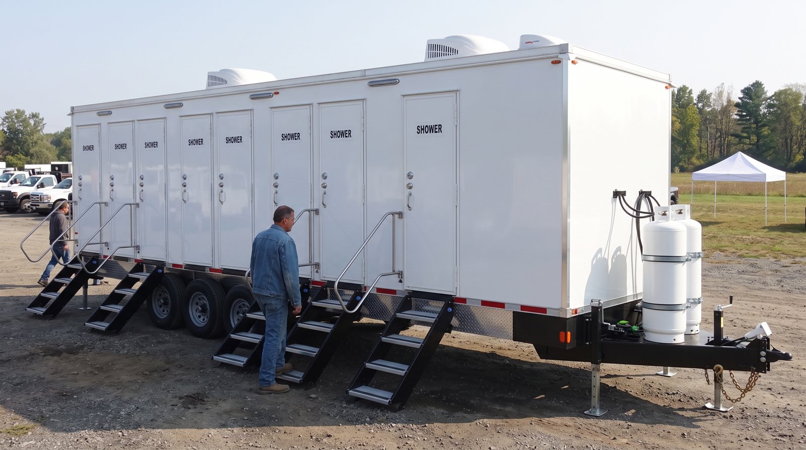 8-station shower trailer in a gravel lot with two crew members, work trucks and a white canopy tent