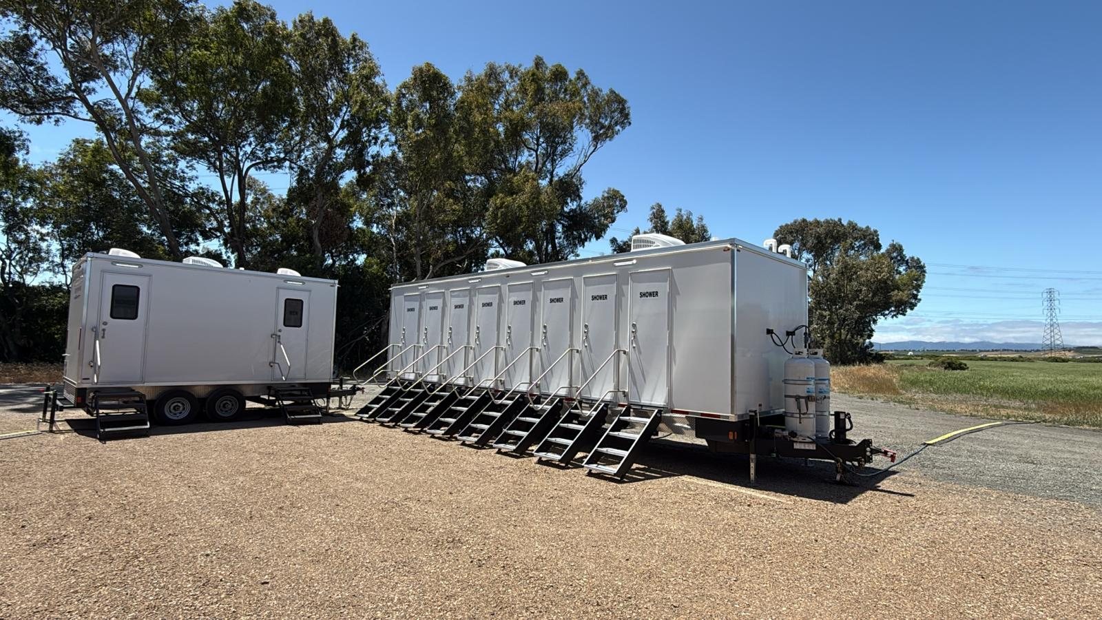 8-station private shower trailer with a stair at every door and propane tanks on the tongue, set on a gravel pad beside an open field, with a 2-station trailer parked next to it